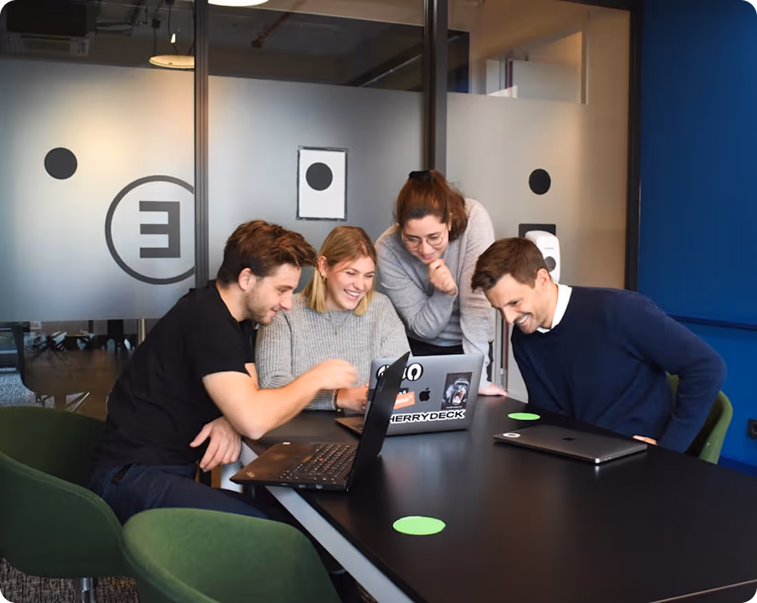 Four young professionals smiling and collaborating around laptops in a modern office meeting room.